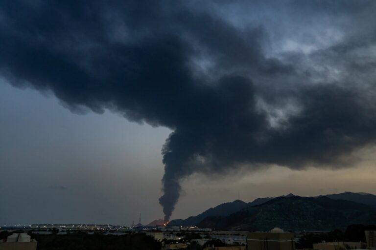 A massive plume of dark smoke rises from a distant fire near mountains behind a coastal industrial area at dusk.