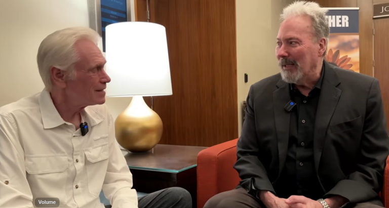 Two men sit facing each other in conversation beside a lit table lamp with a round gold base