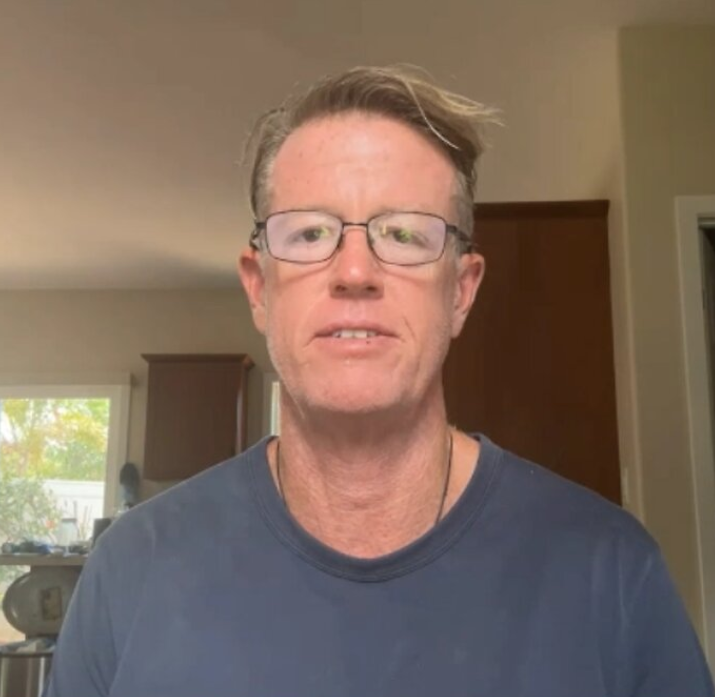 A middle-aged man with glasses and tousled light-brown hair, wearing a blue t-shirt, speaks in a home interior.