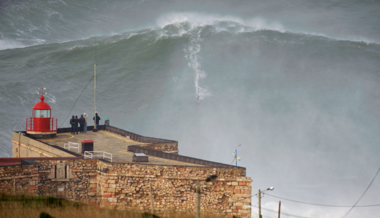 A surfer rides a massive wave behind a stone fort with a red lighthouse, where several people watch from the platform.