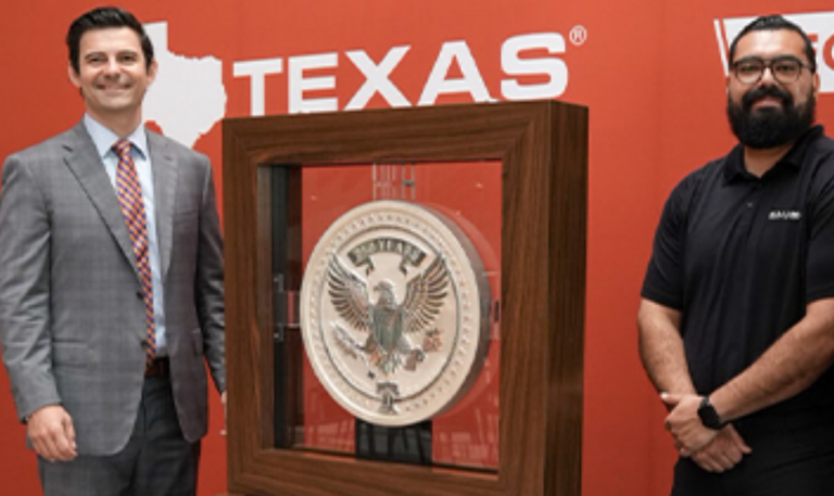 Two men flank a large silver coin with an eagle design in a wooden display case against a red wall with Texas signage