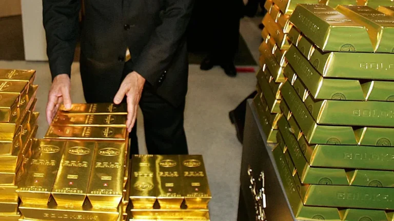 Man in a dark suit standing between tall stacks of gold bars, hands touching the bars