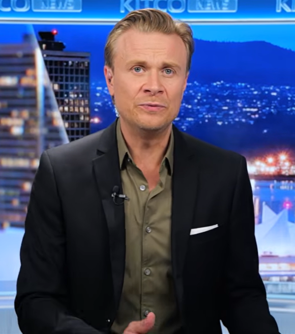 Blond news anchor in a dark blazer and olive shirt, seated in a studio with a nighttime city skyline backdrop