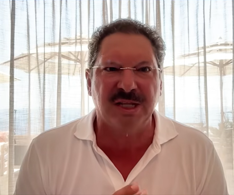 Man with glasses and mustache in a white polo speaking in front of sheer curtains with beach umbrellas behind.